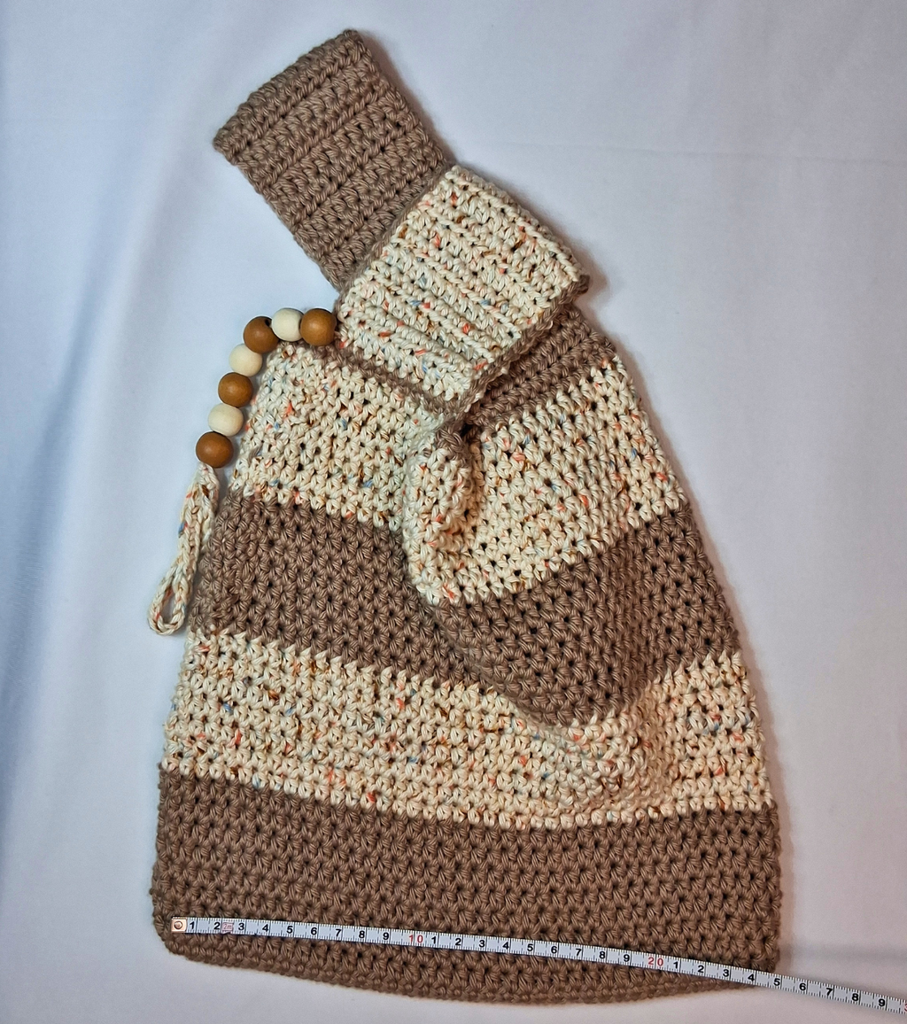 Full view of a medium crochet Japanese knot bag in beige and spunky sand striped 100% cotton laid flat with a measuring tape showing size, wood bead embellishment visible